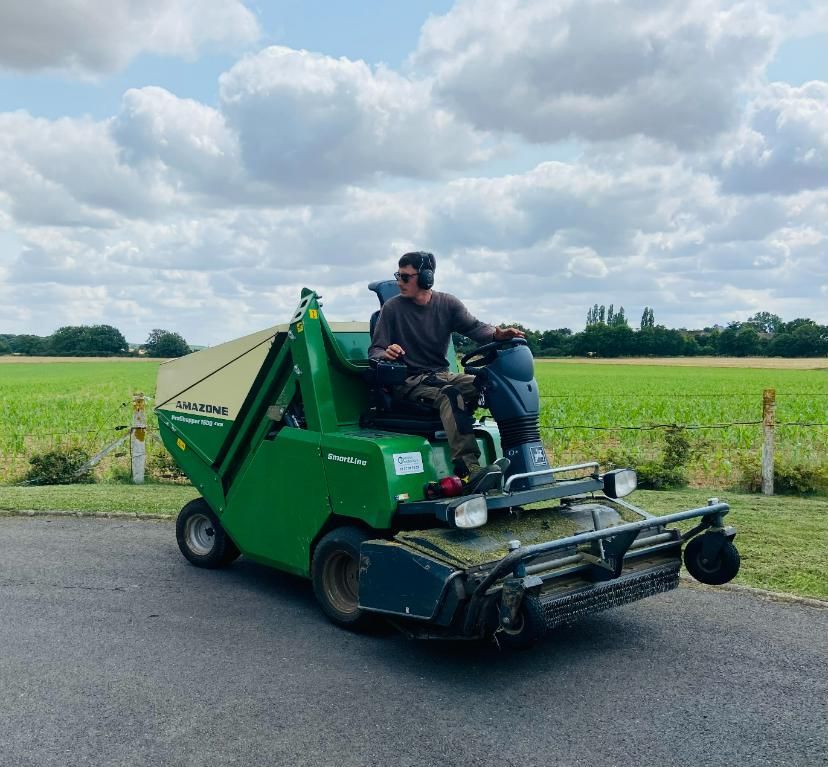 Un homme utilise une tondeuse à gazon verte et beige sur de l'asphalte, avec un champ et le ciel en arrière-plan.