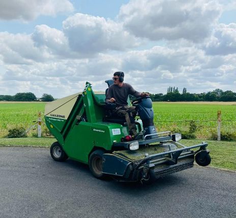 Un homme utilise une tondeuse à gazon verte et beige sur de l'asphalte, avec un champ et le ciel en arrière-plan.