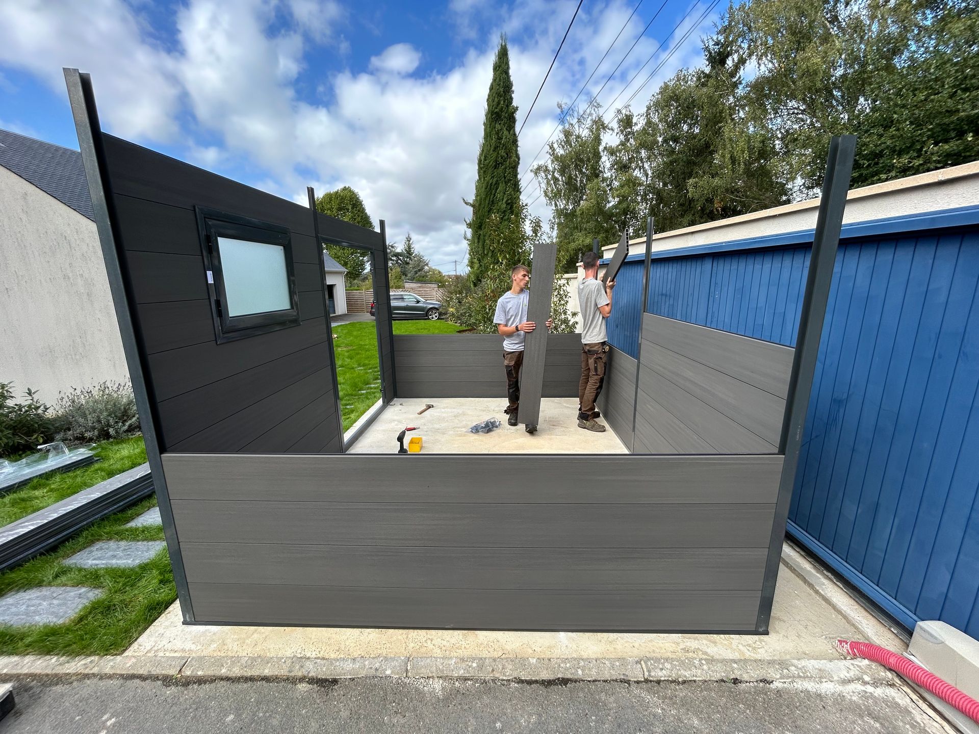 Un abri de jardin vert et une pergola noire dans une cour arrière avec du gazon artificiel et une clôture en bois sous un ciel bleu.