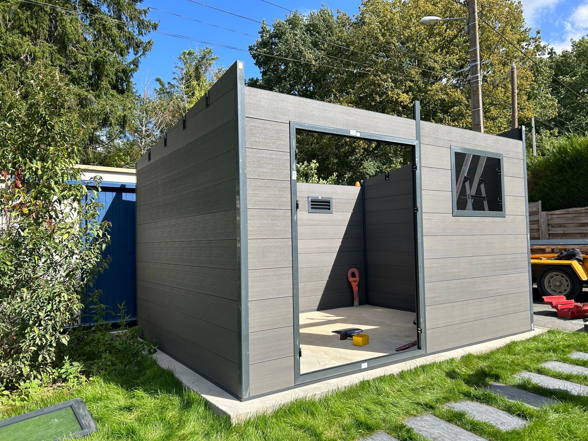 Abri de jardin blanc avec des bordures grises et une porte gris foncé, situé dans une cour herbeuse, partiellement entourée de feuillage vert.