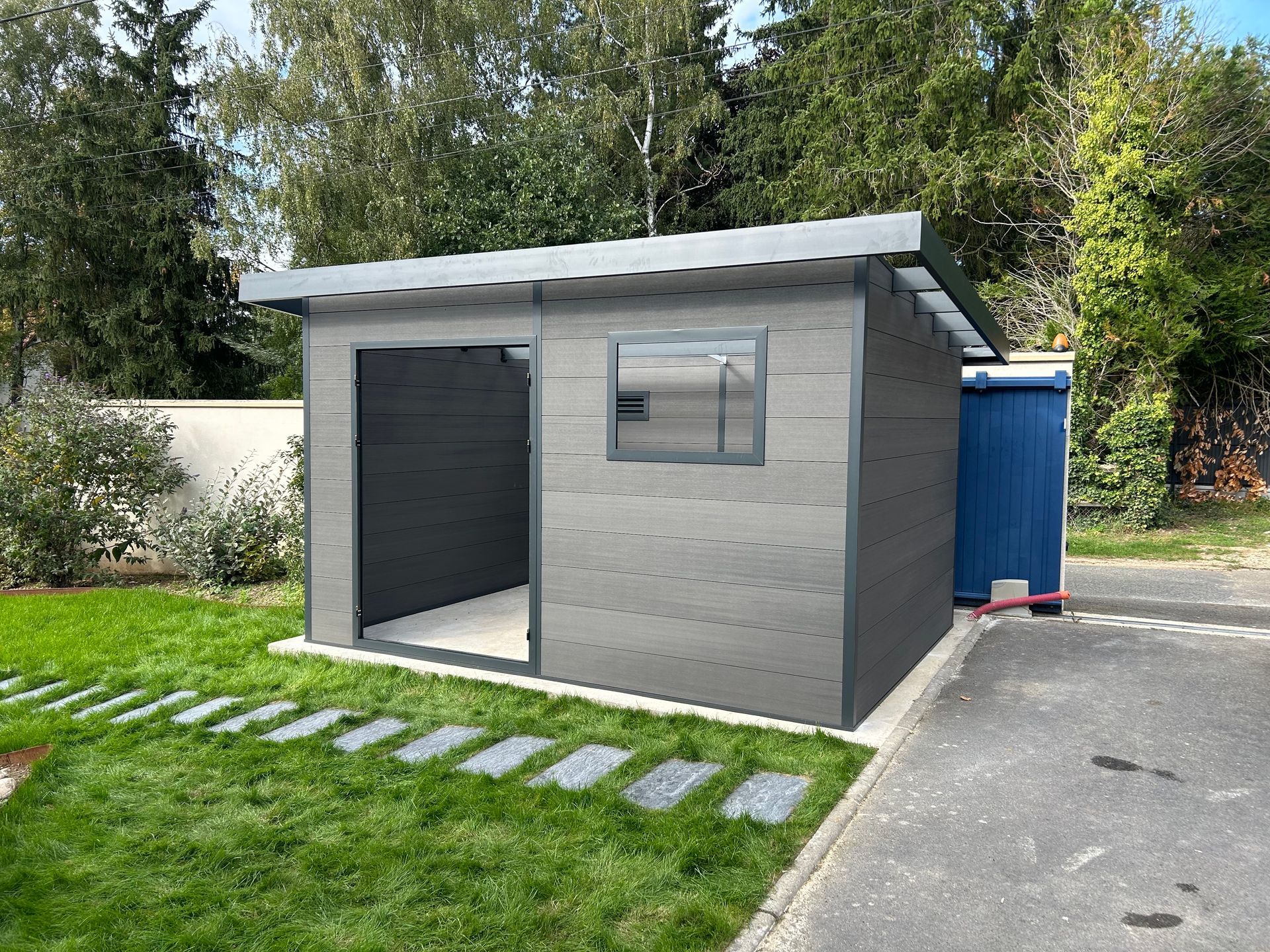 Abri de jardin en bois blanc avec des boiseries grises, une porte sombre et une petite fenêtre, entouré d'herbe verte et d'un arbre.