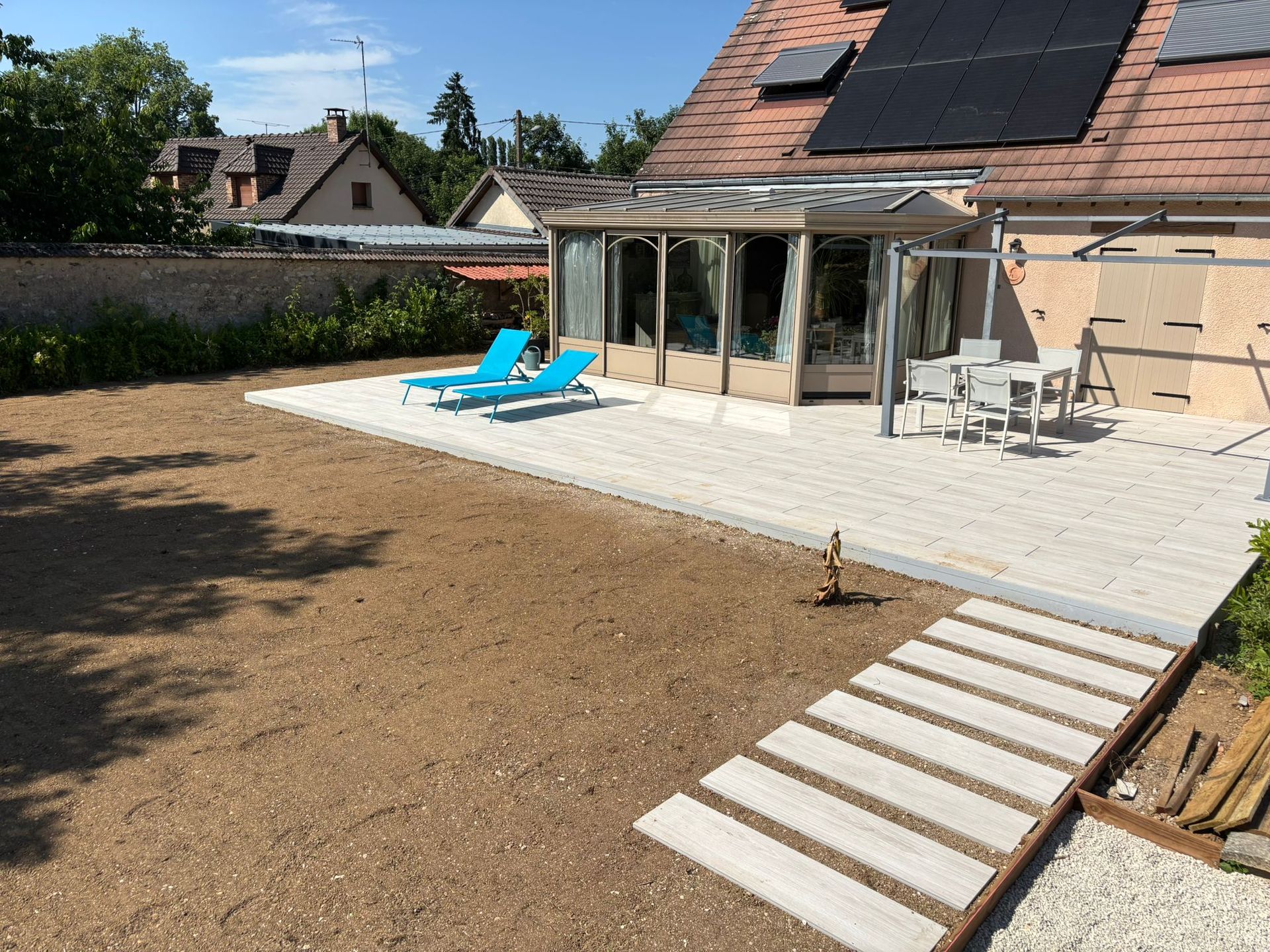 Maison au toit de tuiles rouges, terrasse en bois, coin salon extérieur, volets bleus et pelouse verte sous un ciel bleu azur.