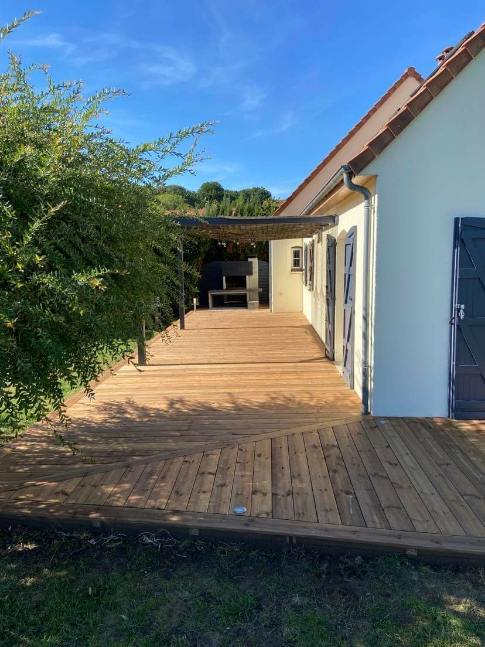 Terrasse en bois longeant une maison blanche avec un patio ombragé, une végétation luxuriante et un ciel bleu.