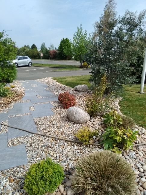 Chemin de pierres traversant un jardin paysager avec des plantes variées et des galets, voiture en arrière-plan.