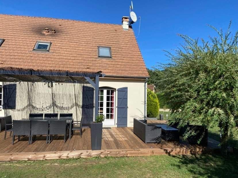 Maison au toit de tuiles rouges, terrasse en bois, coin salon extérieur, volets bleus et pelouse verte sous un ciel bleu azur.