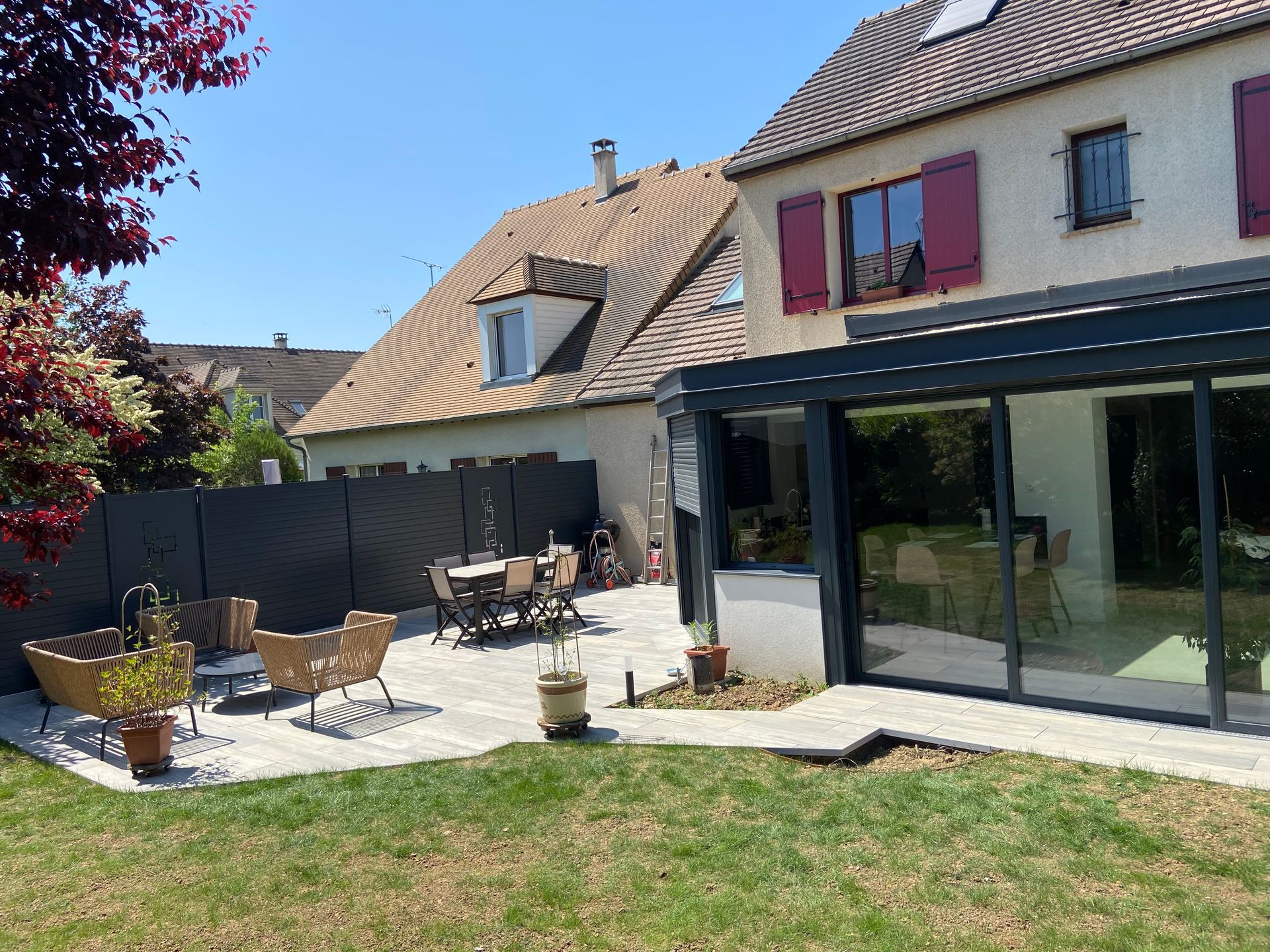 Jardin avec terrasse, pelouse et maison. Table et chaises sur la terrasse. Journée ensoleillée.