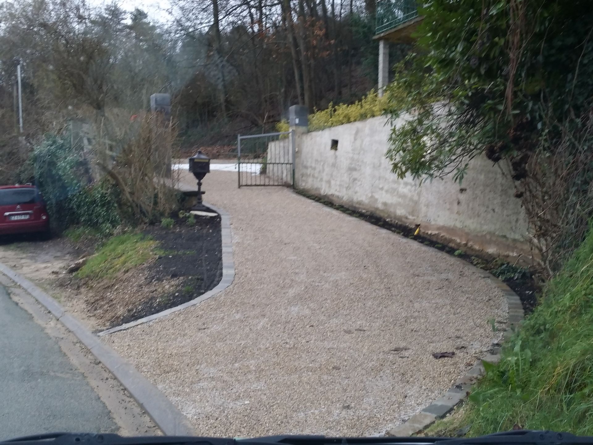 Allée de gravier en pente douce, longeant un muret blanc. Petite voiture rouge garée à gauche.
