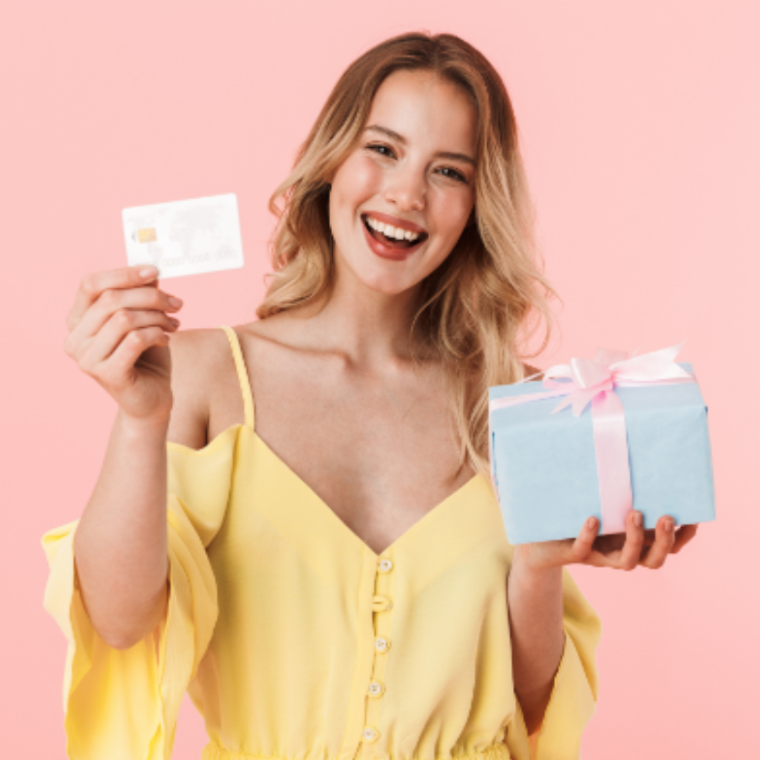 Donna sorridente con un vestito giallo, che tiene in mano una scatola regalo e un biglietto, su uno sfondo rosa