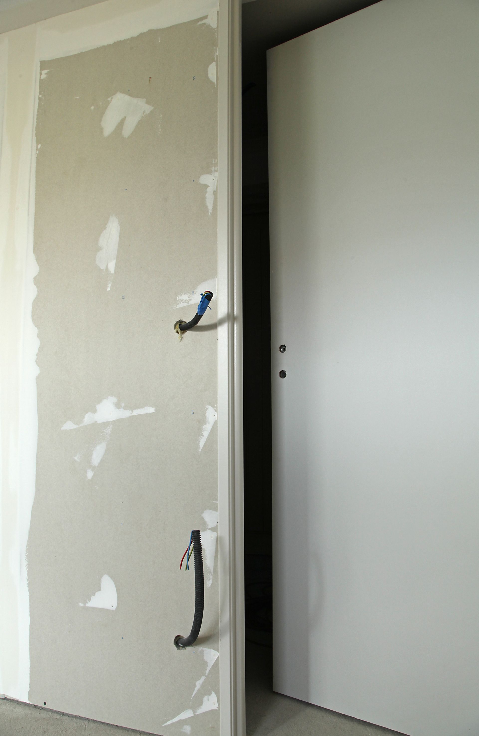 Une porte blanche entrouverte, révélant un mur partiellement fini avec des câbles électriques et une seconde porte blanche fermée.