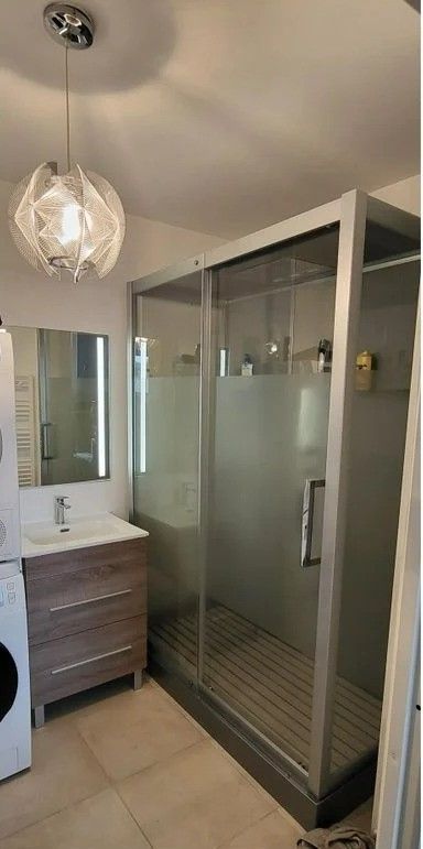 Salle de bains avec douche, meuble-lavabo et suspension lumineuse. La douche est équipée d'une porte en verre dépoli et d'un receveur en bois.