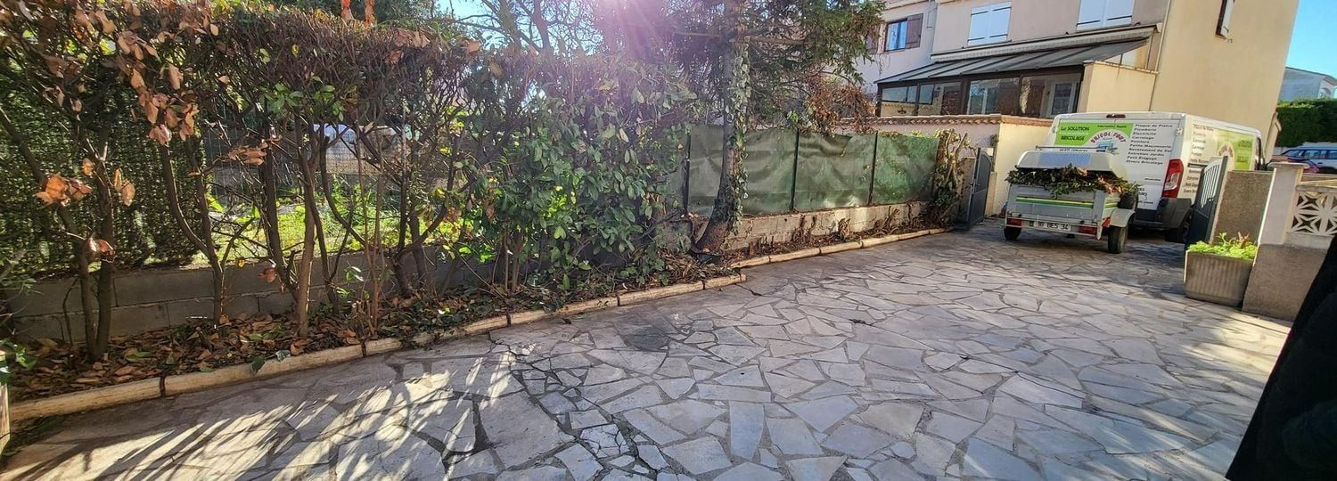 Allée pavée de pierres avec des arbustes verts, une clôture et une camionnette commerciale blanche garée à côté d'un bâtiment.