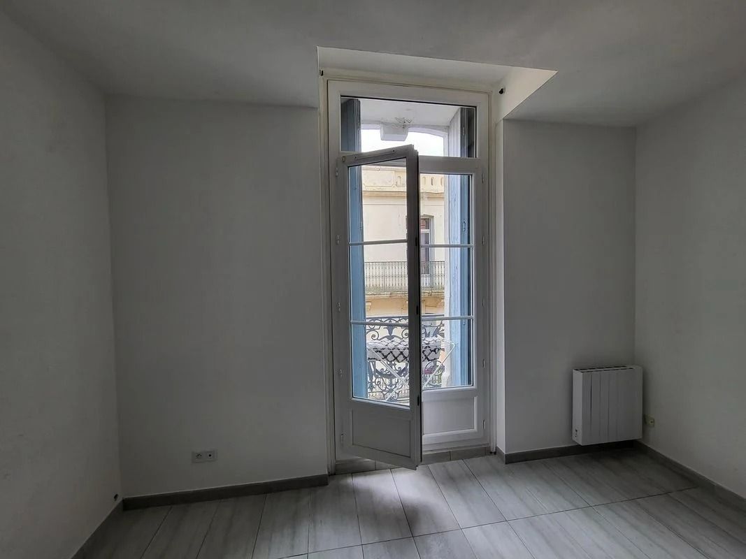 Pièce vide avec une haute fenêtre entrouverte laissant entrer la lumière. Murs blancs, sol gris, radiateur à droite.