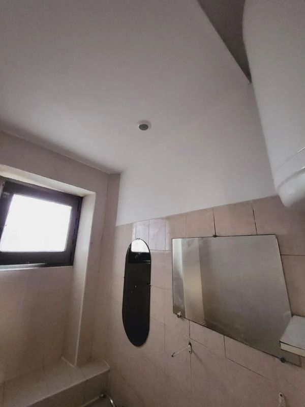 Intérieur de salle de bains avec fenêtre, miroirs et plafonnier. Tons beiges et blancs.