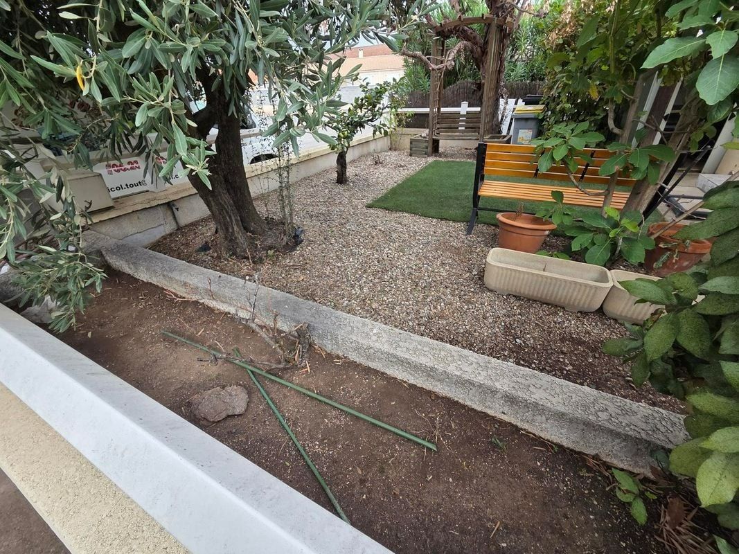 Un petit jardin avec un sol recouvert de gravier, des arbres, du gazon artificiel et un banc en bois.