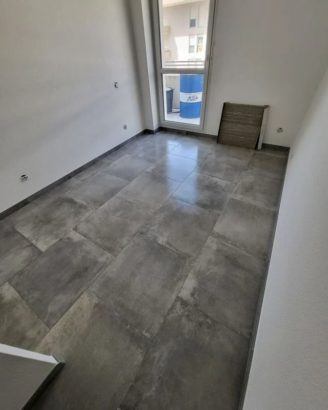 Pièce vide avec sol carrelé gris, murs blancs et une fenêtre laissant apparaître un tonneau bleu à l'extérieur.