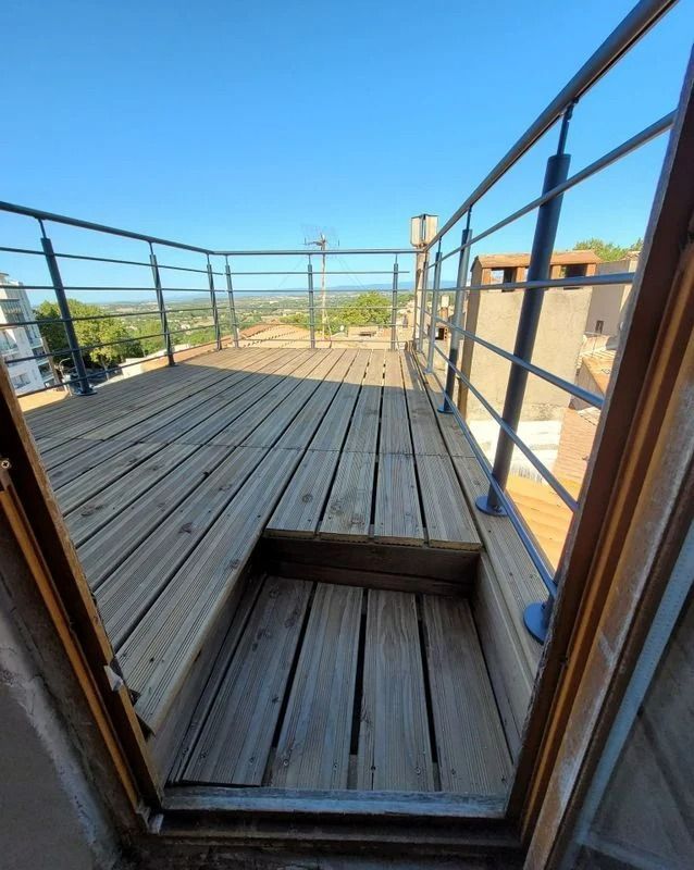 Terrasse en bois avec rambarde en métal, accessible par une porte ouverte, ville en arrière-plan.