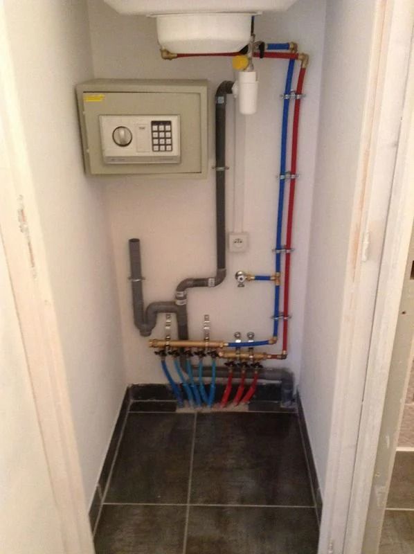 Plomberie dans un petit meuble de salle de bains avec coffre-fort. Tuyaux rouges et bleus contre un mur blanc ; sol carrelé noir.