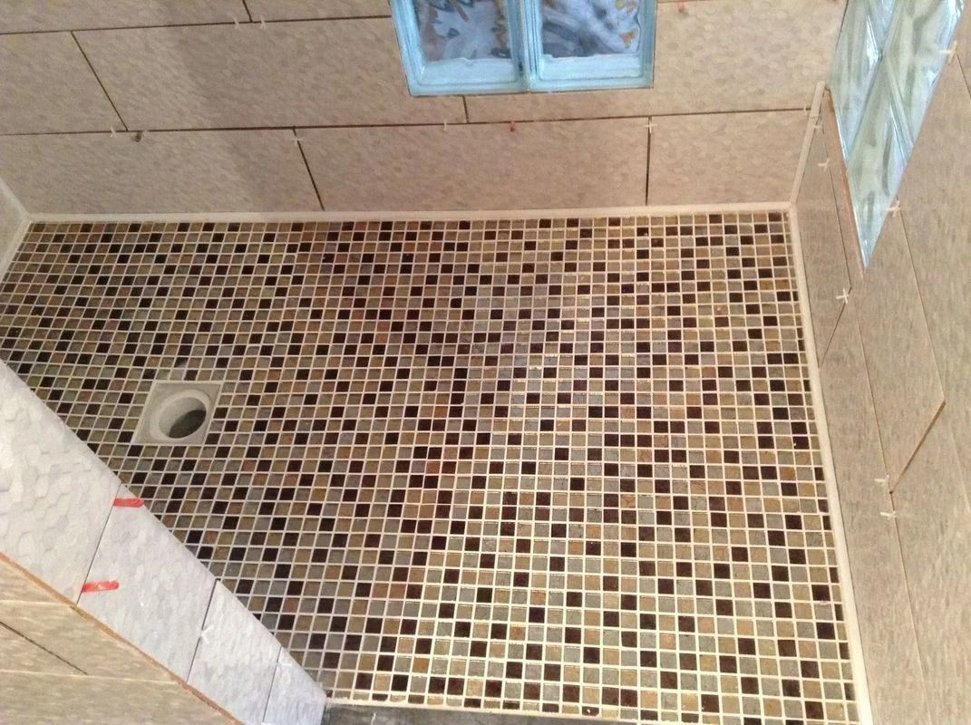 Cabine de douche avec murs et sol en mosaïque, couleurs neutres. Une bonde est visible.