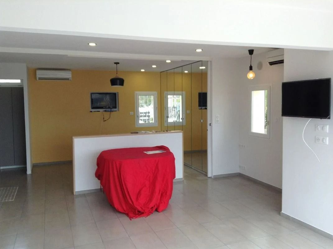 Espace intérieur vide avec des murs blancs et jaunes, une table recouverte d'une nappe rouge et deux téléviseurs.