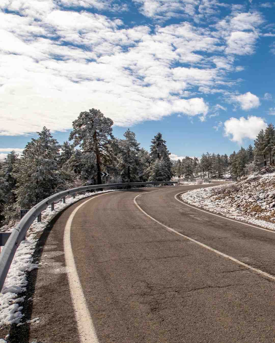 Carretera sinuosa a través de un paisaje nevado, bordeada de árboles cubiertos de nieve, bajo un cielo azul con nubes.