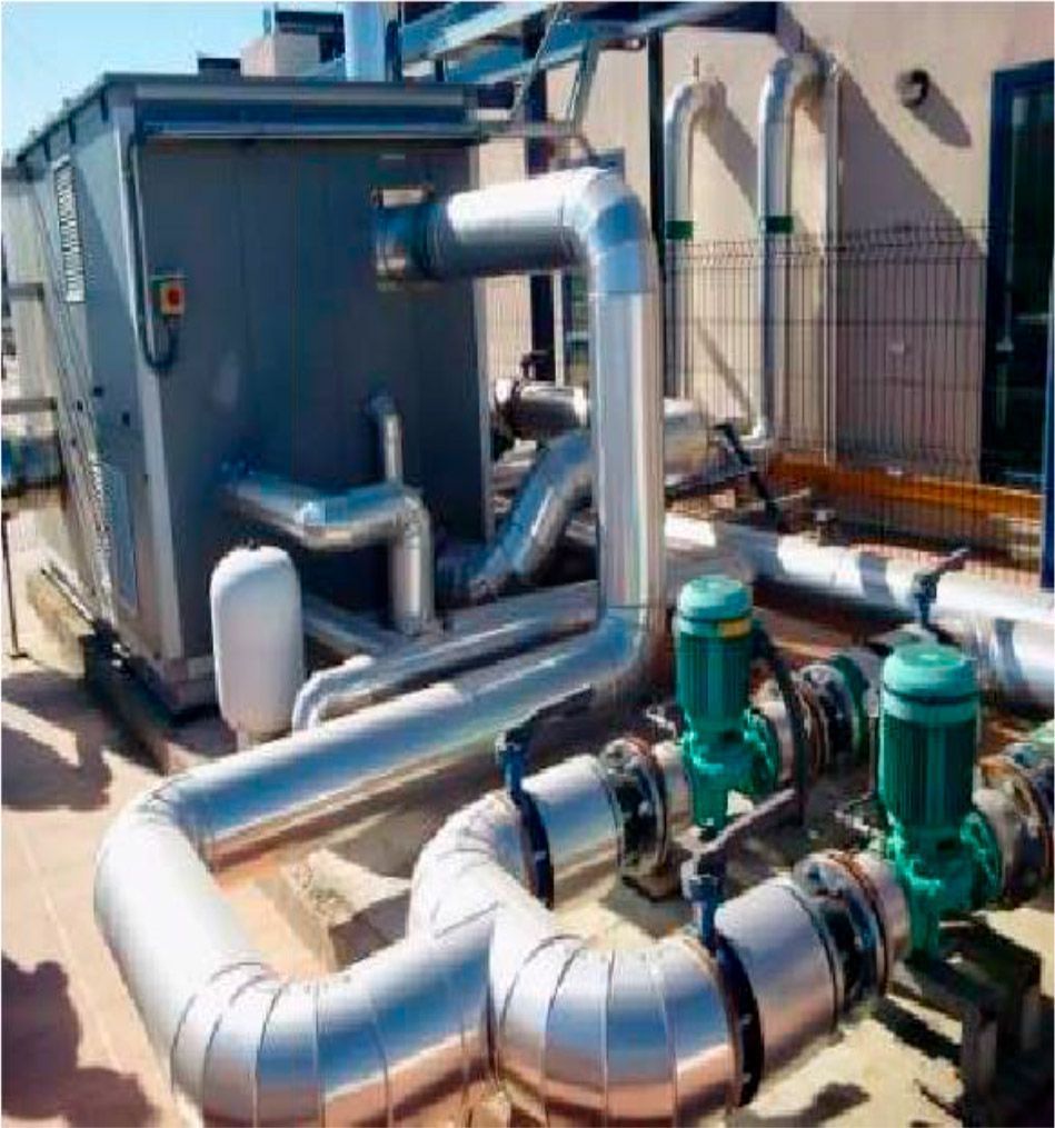 Equipo de climatización (HVAC) en una azotea: unidad gris grande, tuberías aisladas, dos bombas verdes y una valla metálica.