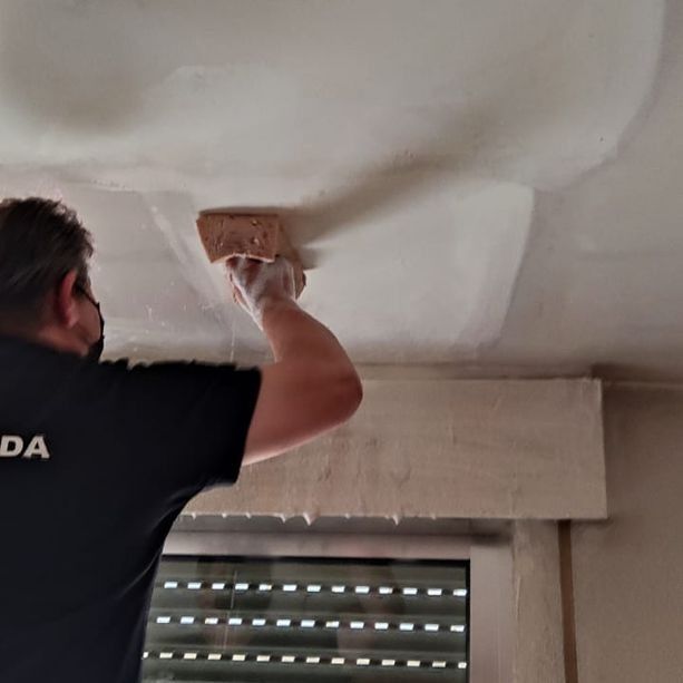 Persona alisando yeso en un techo con una herramienta similar a una esponja, cerca de una ventana con la persiana cerrada.