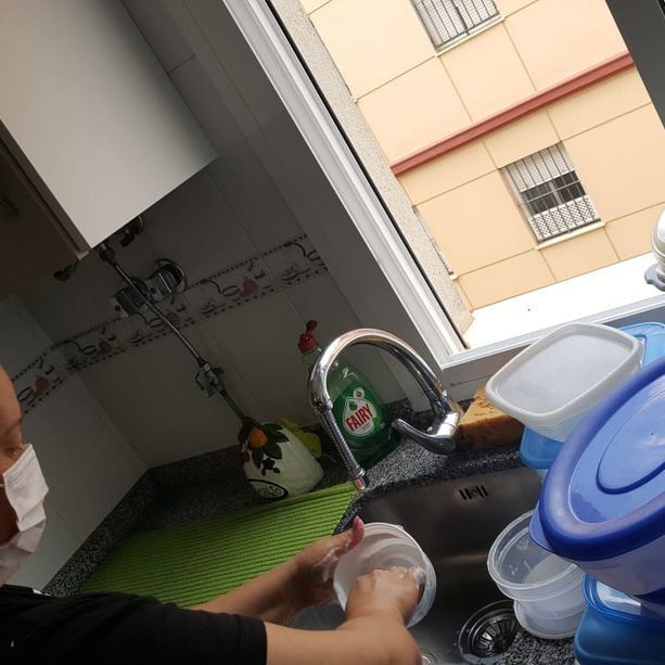 Persona que lleva una mascarilla lavando un recipiente transparente en el fregadero de la cocina.