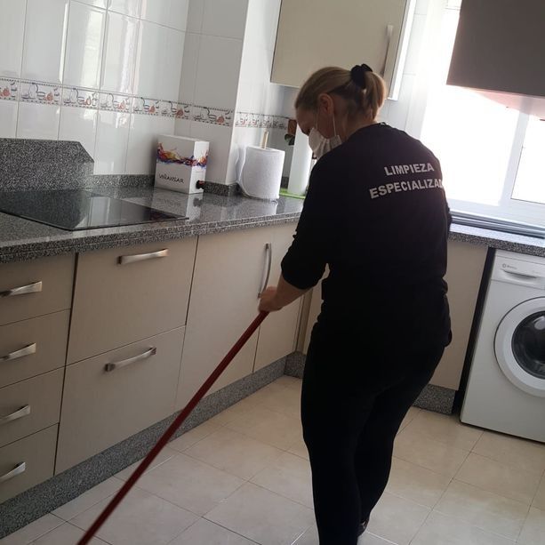 Mujer fregando el suelo de la cocina. Lleva mascarilla y uniforme negro con la palabra 