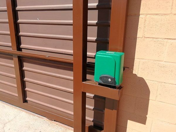 Puerta corrediza de metal marrón con abridor automático verde adosada a una pared de ladrillo marrón claro.