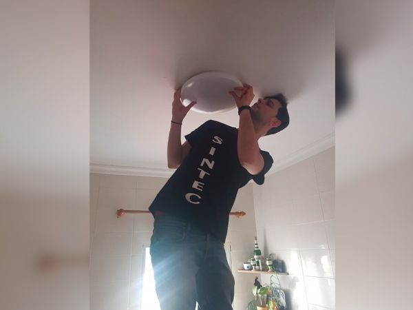 Hombre instalando una lámpara de techo. Está subido a un taburete, estirando la mano hacia arriba. La habitación tiene paredes blancas y una estantería sencilla.