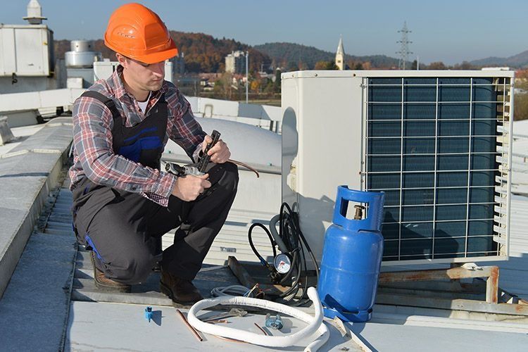 Un técnico de HVAC se arrodilla en el techo, trabajando en una unidad de aire acondicionado; hay un tanque de propano azul cerca.