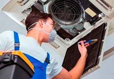 Técnico de HVAC limpiando un filtro de aire con un cepillo, usando una máscara, trabajando en una unidad de techo.