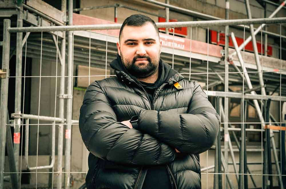 Mann in schwarzer Daunenjacke steht mit verschränkten Armen vor einem Metallzaun und einem Baugerüst.