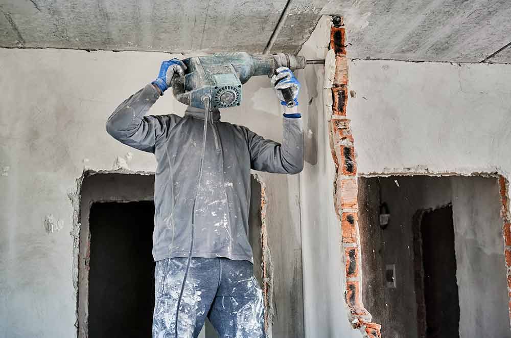 Ein Arbeiter bricht mit einem Presslufthammer Beton um ein senkrechtes Rohr in einem unfertigen Raum auf.