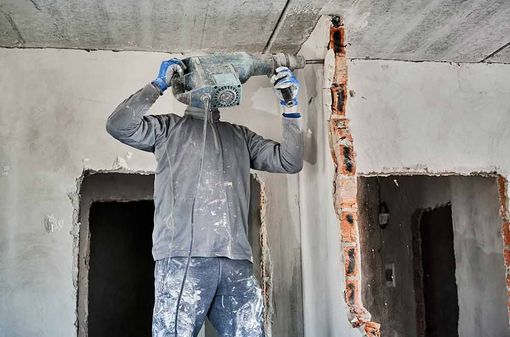 Ein Arbeiter bohrt in einer Betonwand im Innenbereich, daneben liegen freiliegende vertikale Rohrleitungen und zwei Türöffnungen.