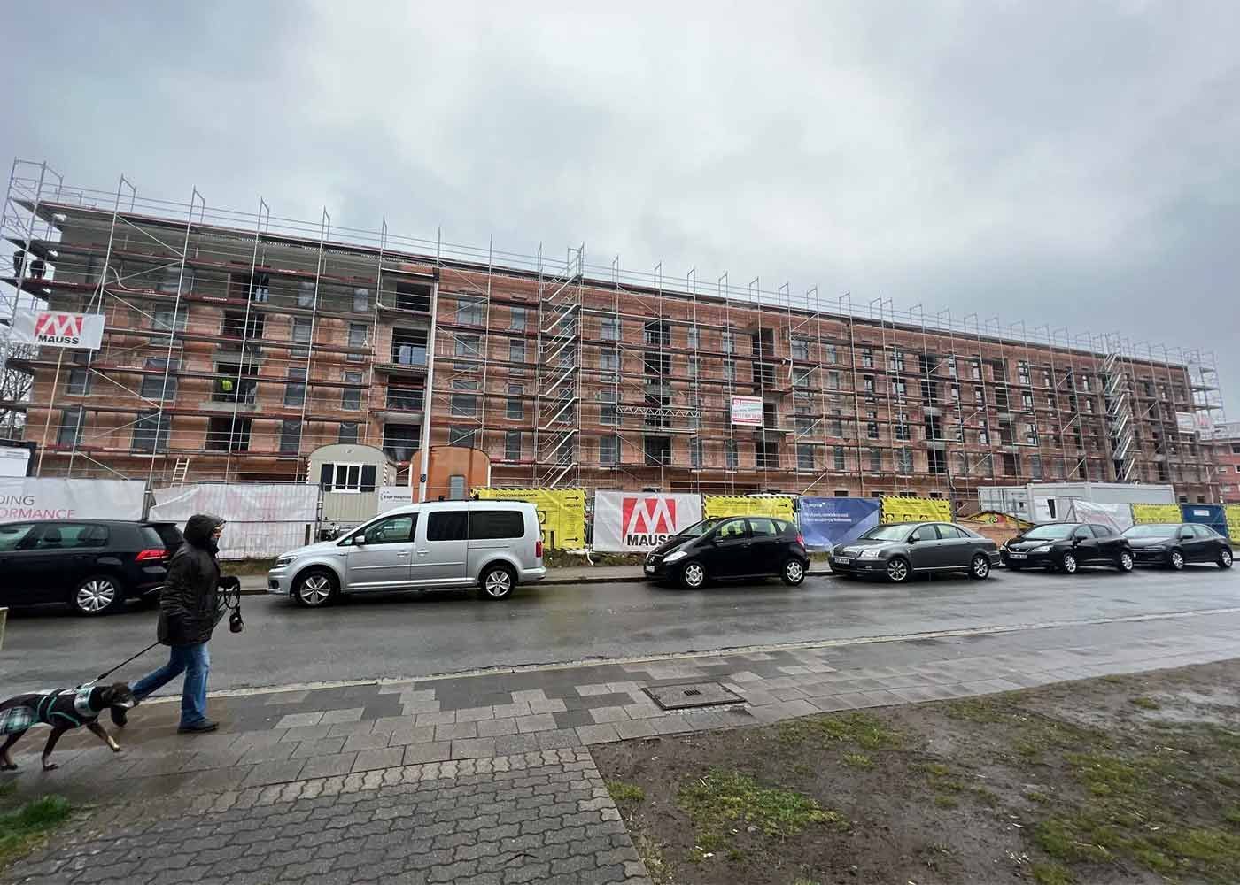 Ein langes, im Bau befindliches Backstein-Mehrfamilienhaus steht an einer regnerischen Straße mit parkenden Autos und einem Fußgänger.