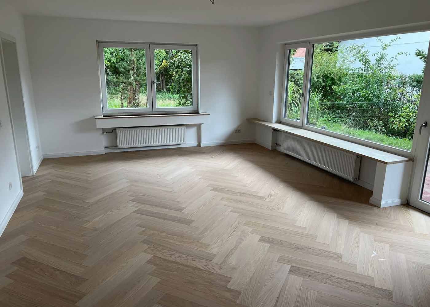 Heller, leerer Raum mit Fischgrätparkett, großen Fenstern und Heizkörpern unter den Fensterbänken.