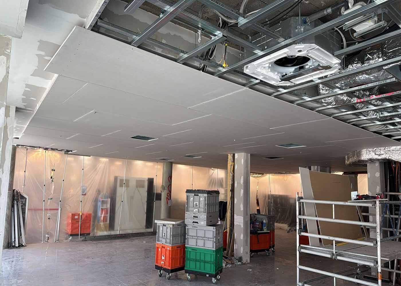 Innenansicht eines im Bau befindlichen Gebäudes mit Gipskartonwänden, freiliegender Decke und gestapelten Materialien auf dem Boden.