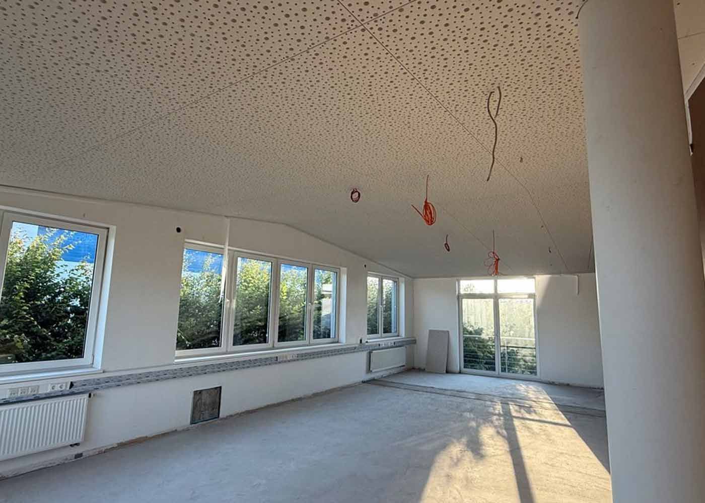 Leeres, unfertiges Wohnzimmer mit weißen Wänden, großen Fenstern und Sonnenlicht auf dem Boden.