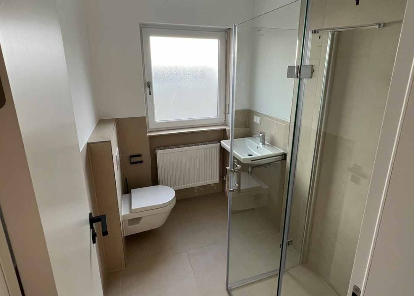 Kleines Badezimmer mit Toilette, Waschbecken, Dusche und Fenster in neutralen Beige- und Weißtönen.