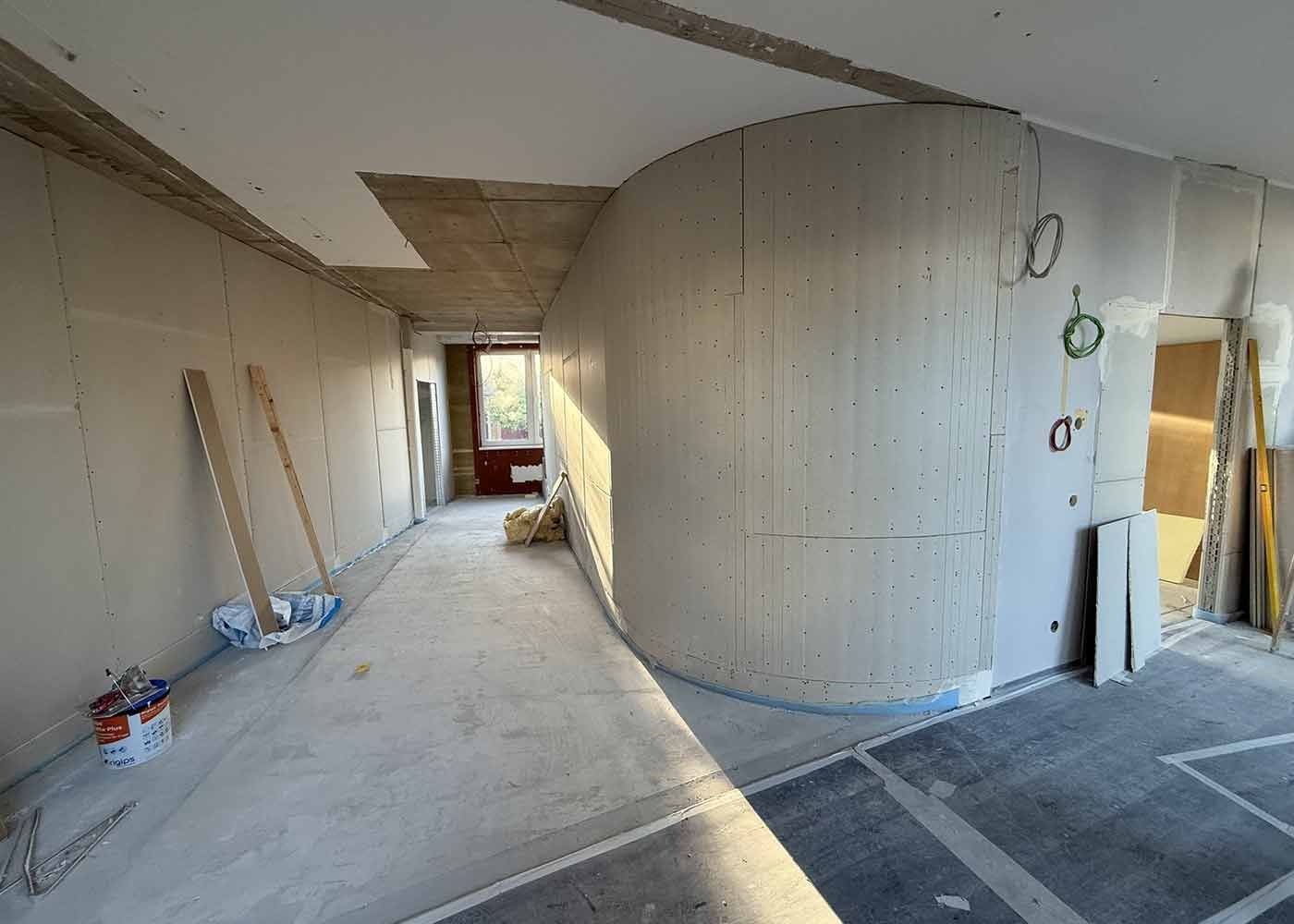 Unfertiger Innenflur mit gebogener Gipskartonwand, freiliegenden Werkzeugen und Baumaterialien auf dem Boden.