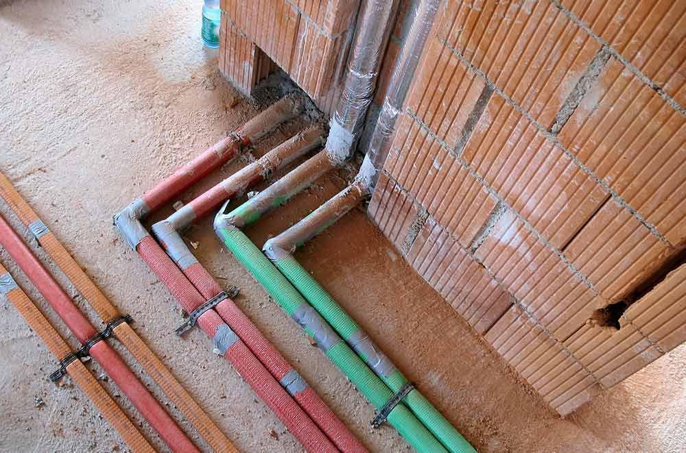 Farbige Rohre verlaufen entlang einer Mauer auf einer Baustelle, darunter rote, grüne und graue Leitungen.