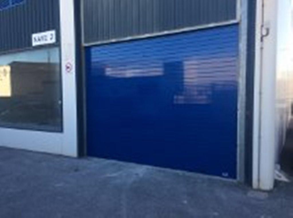 Puerta enrollable industrial azul en un edificio, con una ventana y un cartel cerca.