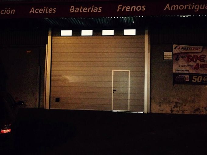 Exterior de un garaje por la noche; gran puerta superior con una puerta más pequeña, iluminada por luces de techo.