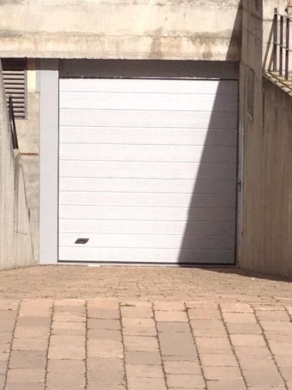 Puerta de garaje blanca con marco gris, situada entre paredes de hormigón y pavimento de ladrillo.