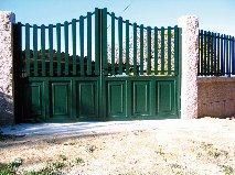 Puerta metálica de color verde con barrotes verticales y paneles rectangulares, engastados en postes de piedra.