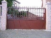 Puerta de entrada de metal marrón con herrajes decorativos, colocada entre pilares de piedra, sobre un camino de entrada pavimentado con ladrillos.