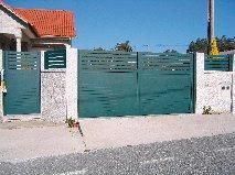 Puerta metálica verde con lamas horizontales, fijada a pilares de hormigón, contra un cielo azul.