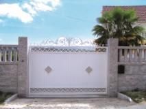 Entrada con portón blanco a una casa con detalles decorativos, flanqueada por pilares de piedra y una valla a juego, bajo un cielo azul.