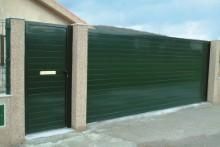 Puerta de entrada de metal verde con puerta lateral, pilares de color canela y cielo azul claro.